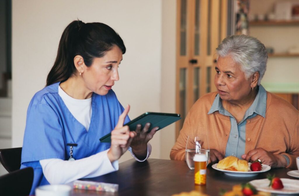 A nurse holding a tablet sits with an older adult during breakfast to discuss their specific dietary needs and medications