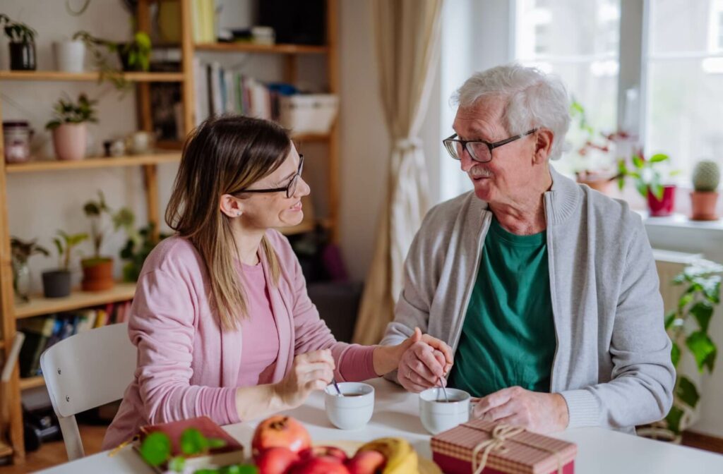 Adult and elderly parent sitting at a table smiling and talking while holding coffee mugs in a cozy plant-filled room