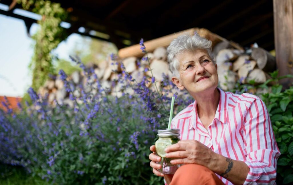 A senior sits in a flower garden and sips on a glass of water at their assisted living home.