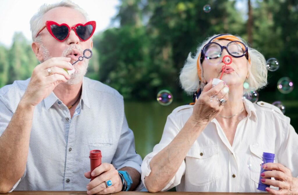 A senior couple wearing fun glasses enjoy blowing bubbles outdoors as an activity that brings them joy