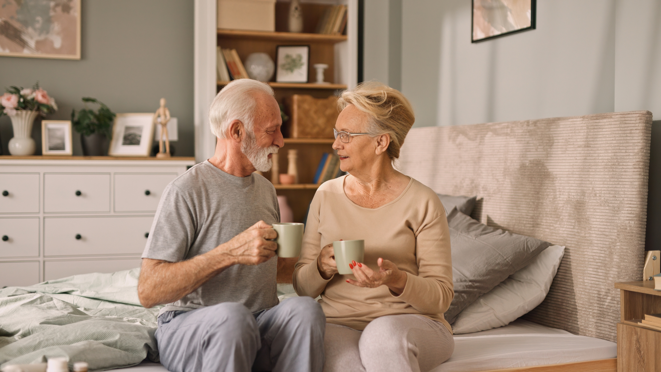 a senior couple sitting on a bed and chatting while drinking coffee