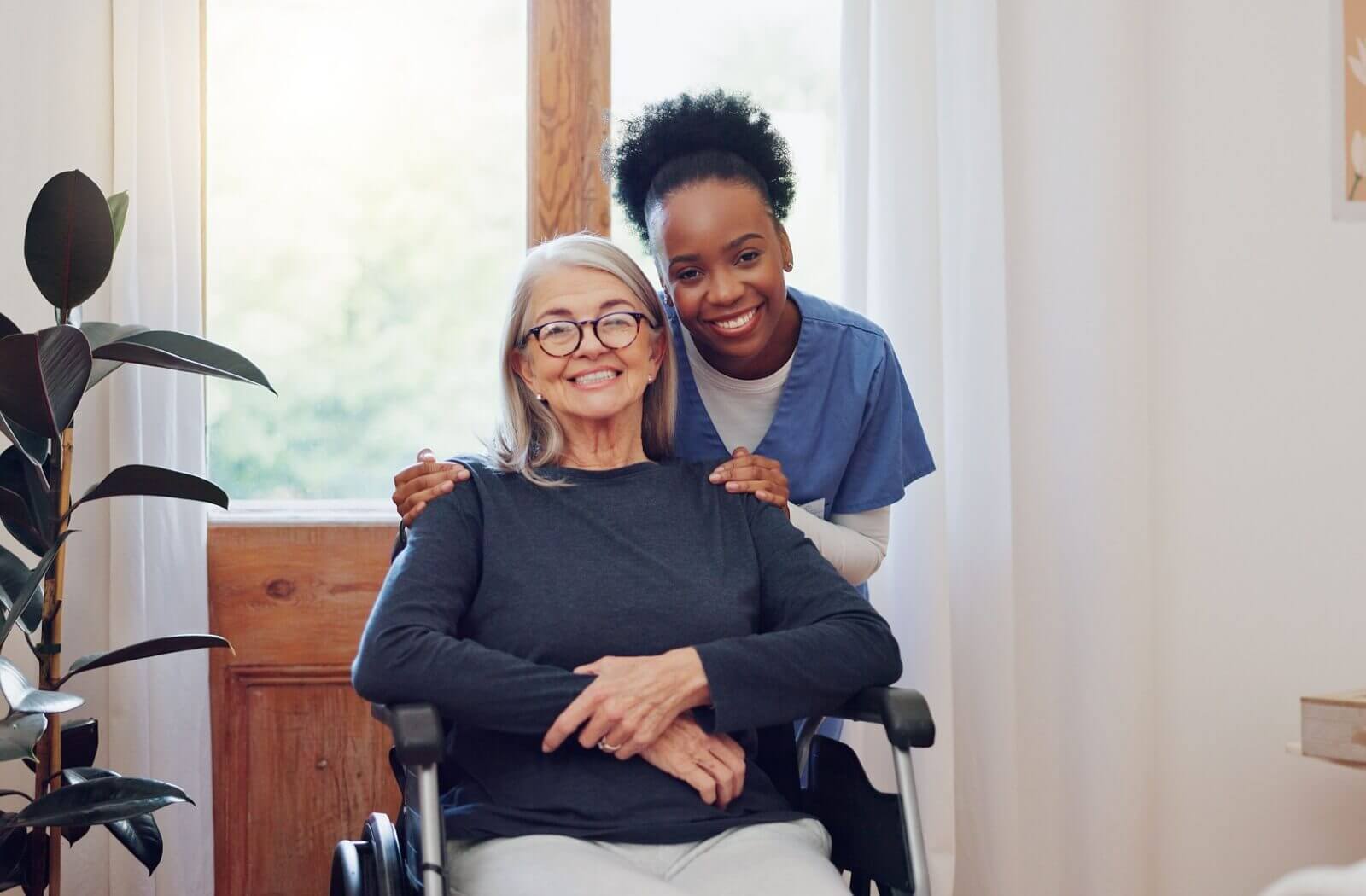 a female healthcare worker smiling and huging an older women in wheelchair in assisted living