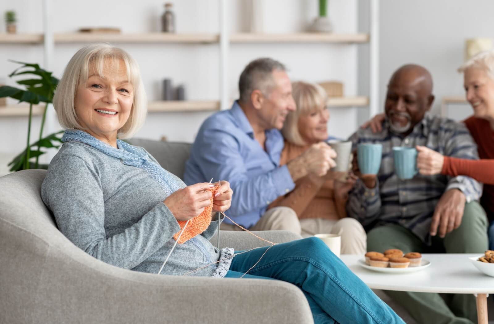 A smiling senior comfortably knits in a chair while friends sit behind them, conversing over coffee and muffins