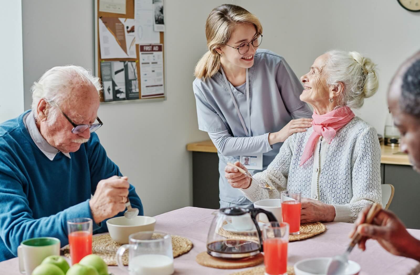 Caregiver interacting with two older adults at a breakfast table with coffee pot and fruits in a bright setting