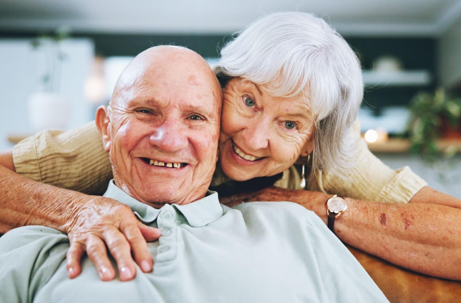 A smiling older adult couple in their senior living home, with one senior hugging the other from behind the couch.