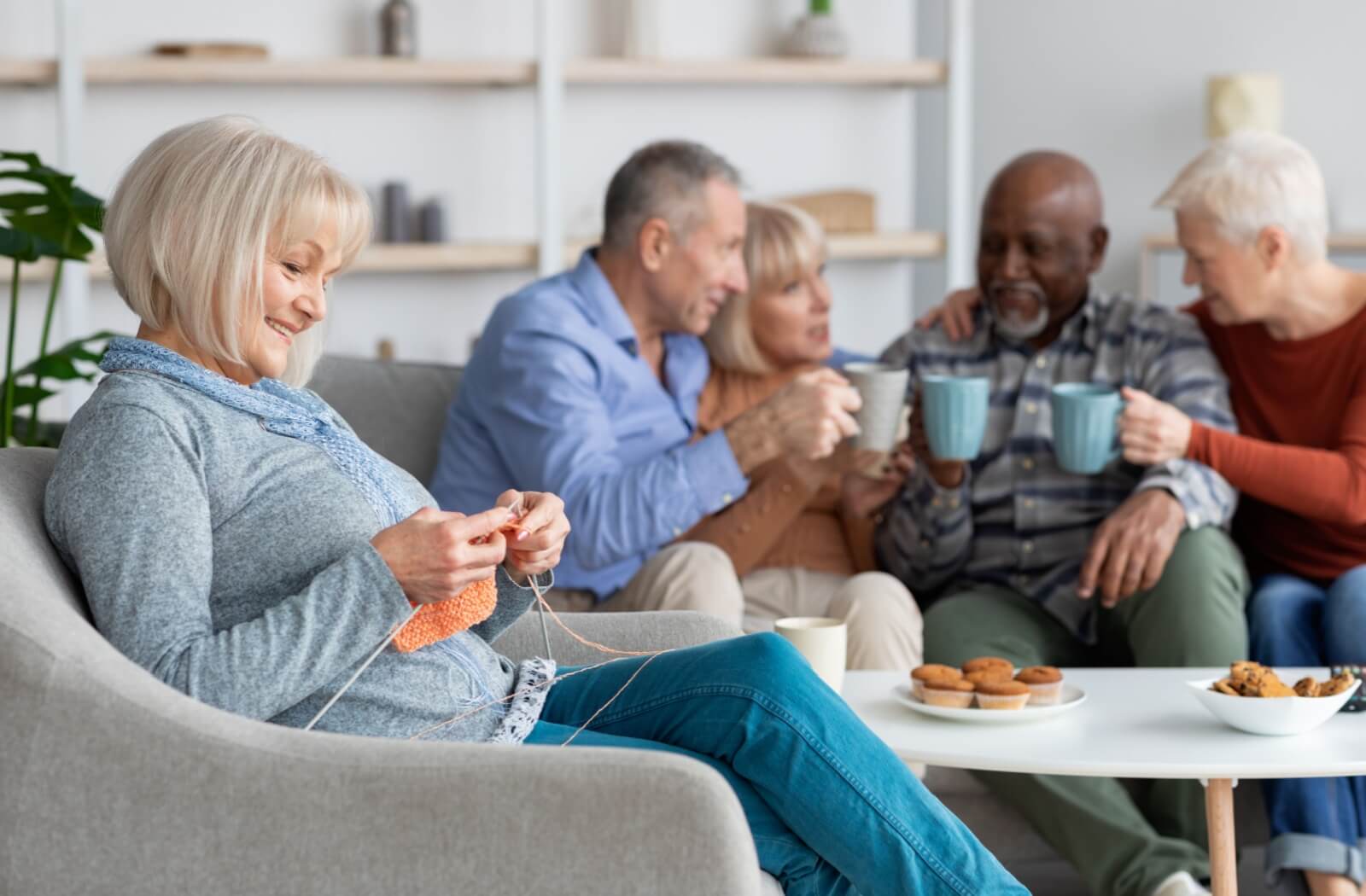 Person knitting on a couch while others enjoy drinks and conversation in a cozy shared space
