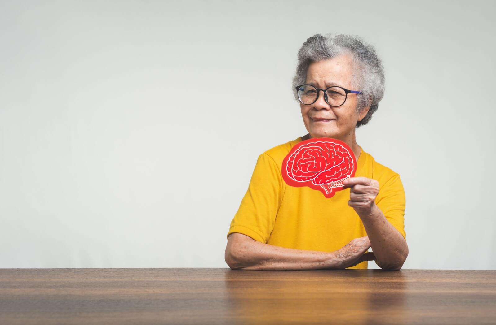 A senior at a wooden table holds up a red cutout image of a brain, symbolizing the support needed for senior mental health