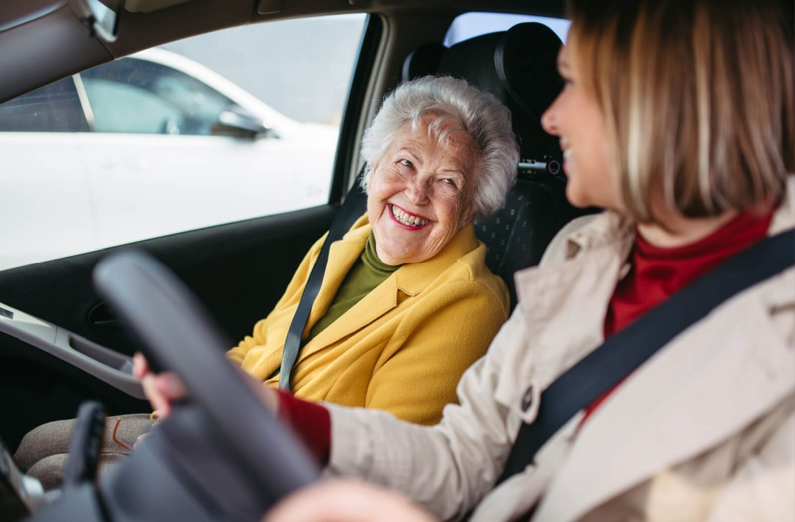 A young adult smiles at their grandparent in the passenger seat of their car, picking them up from senior living for the day