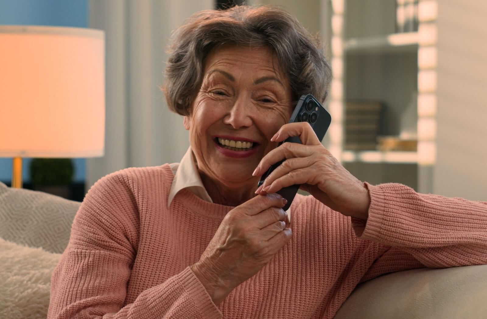 A smiling senior with dementia holds a cellphone with two hands, and is delighted to be talking to a family member on the other side.