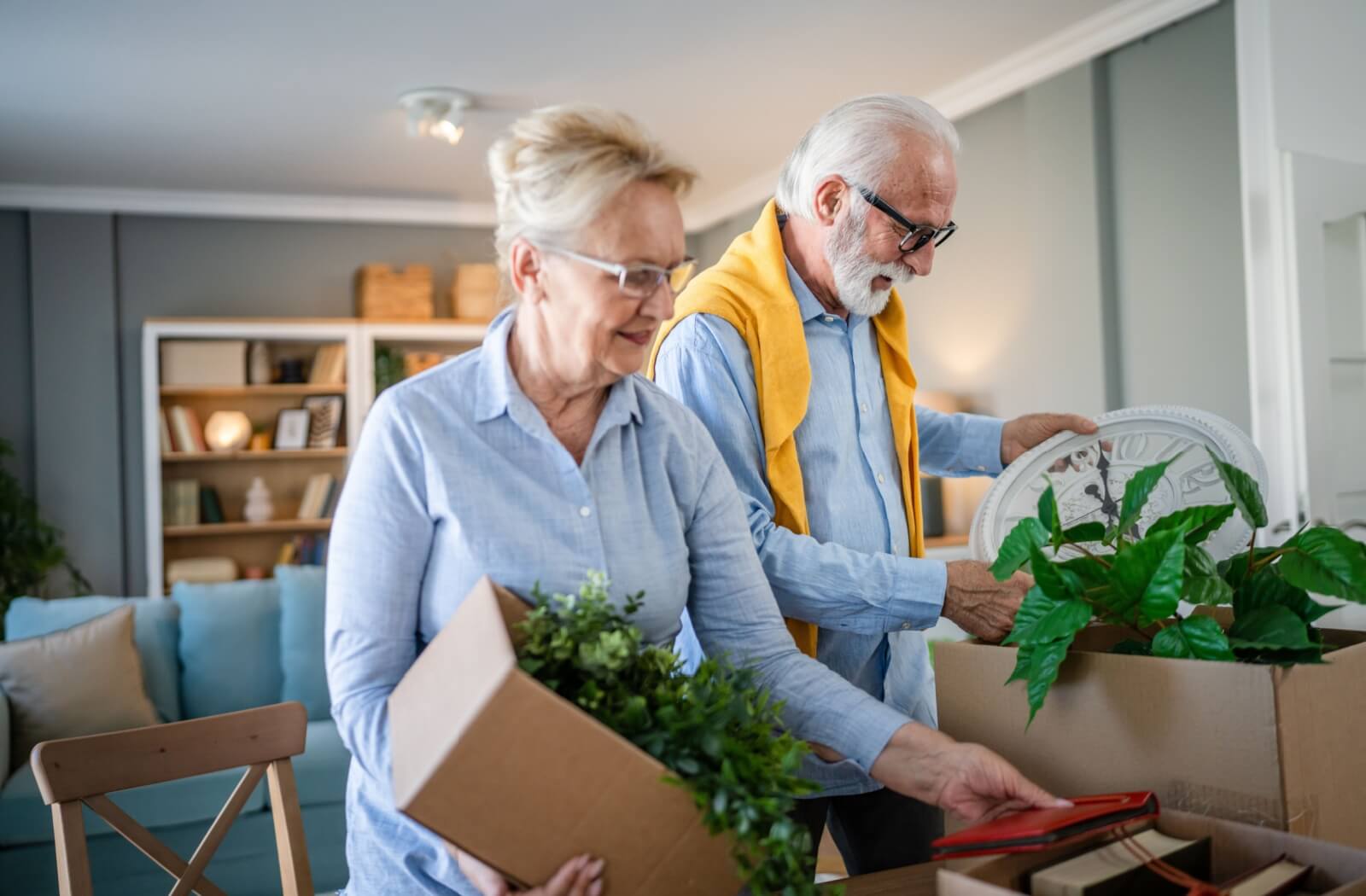 A senior couple is packing boxes with plants and photos because they are moving into senior living.
