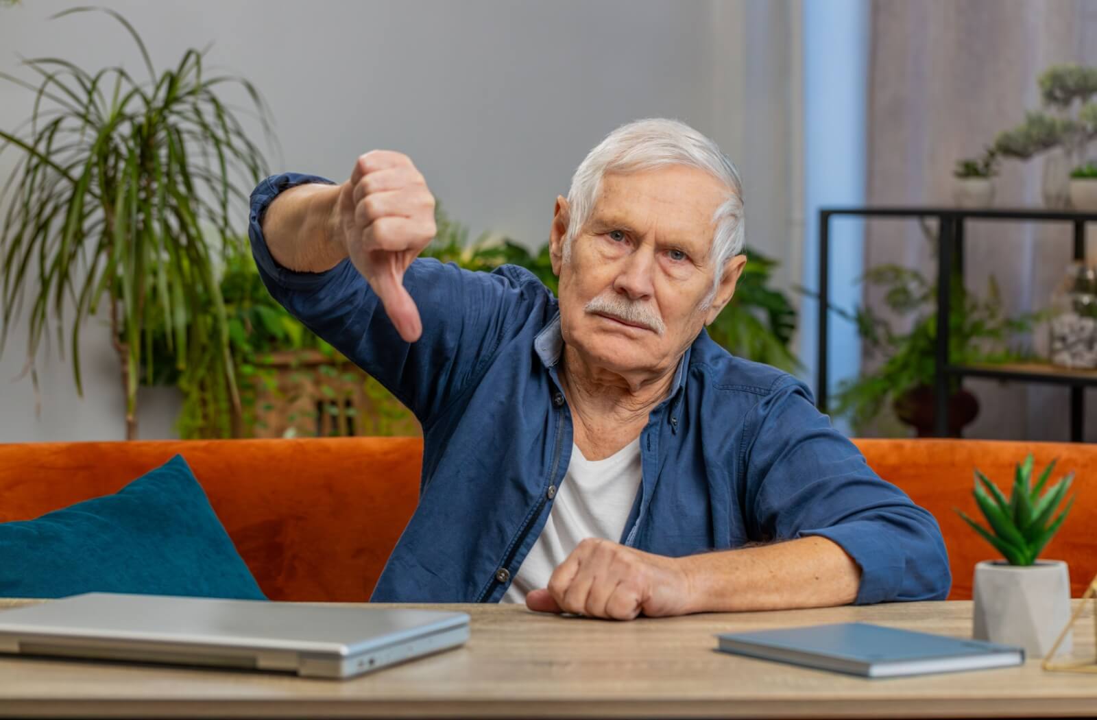 A senior sitting on a bright orange couch at a wooden table gives a thumbs-down sign to express disappointment