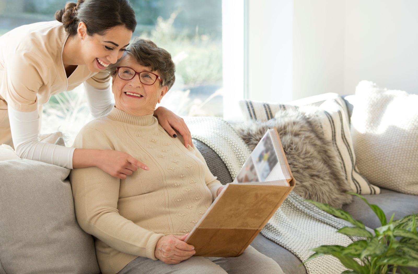 A senior is sitting on a couch in a sunny room, looking at a photo album and sharing fond memories with a caregiver.