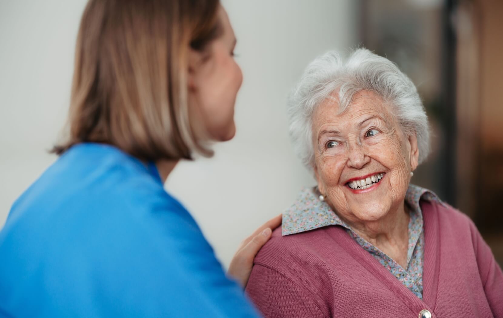 A senior in memory care smiles and jokes with a staff member who places a gentle hand on the resident's shoulder.